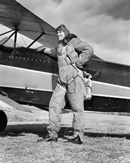 Expressions Prints: 1940s PORTRAIT SMILING MAN AVIATOR PILOT STANDING BY BIPLANE WEARING CANVAS FLYING SUIT HELMET GOGGLES AND PARACHUTE