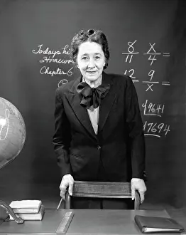 Story Prints: 1940S Smiling School Teacher Standing Behind Her Desk & In Front Of The Black Board With Fractions