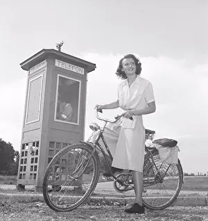 Bicycle Prints: 1940s woman on a bicycle. A smiling young woman on a womens bicycle beside a phone booth