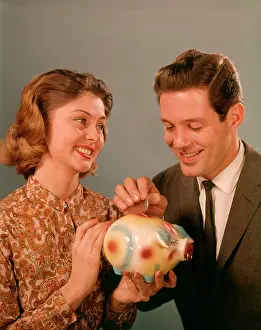 People Adults Prints: 1950S 1960S Happy Smiling Couple Man Woman Putting Coins Into Savings Piggy Bank