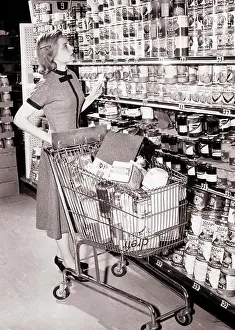 Cart Prints: 1950s 1960s WOMAN PUSHING SHOPPING CART IN GROCERY STORE HOLDING HER SHOPPING LIST AND LOOKING UP AT SHELVES OF CANNED