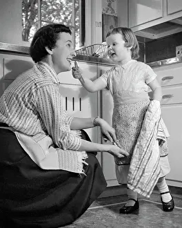 Mothers Day Prints: 1950s LITTLE GIRL AND MOTHER IN KITCHEN BOTH WEARING APRONS GIRL HOLDING DISH TOWEL GESTURING TO MOTHER