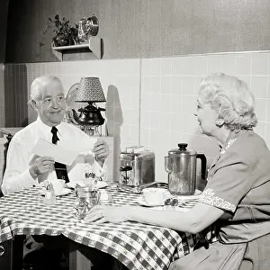 Opening Prints: 1950s SENIOR COUPLE SITTING AT KITCHEN TABLE HAVING TOAST AND COFFEE MAN OPENING MORNING MAIL SMILING HOLDING CHECK