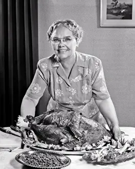 Grandma Prints: 1950s SENIOR WOMAN LOOKING AT CAMERA PLACING LARGE THANKSGIVING TURKEY ON THE DINING TABLE WITH BOWL PEAS AND CELERY