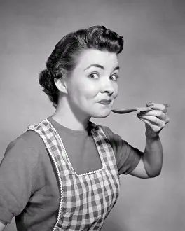 Expressions Prints: 1950s SMILING WOMAN HOUSEWIFE WEARING CHECKERED APRON LOOKING AT CAMERA WITH RAISED EYEBROWS TASTING FROM WOODEN