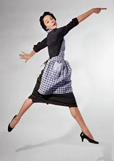Grownups Prints: 1950s WOMAN HOUSEWIFE IN GINGHAM APRON RUNNING AND JUMPING INTO AIR SMILING AND POINTING INDEX FINGER LOOKING AT CAMERA