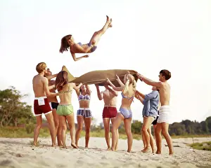 Suits Prints: 1960s 1970s BEACH GAMES GROUP OF TEENAGE BOYS AND GIRLS WEARING BATHING SUITS BEACH BLANKET TOSSING ONE GIRL INTO THE