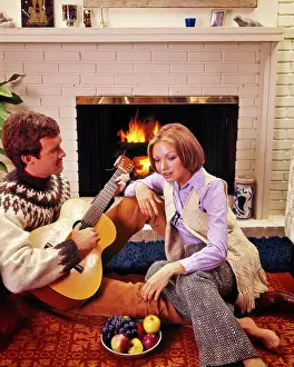 Husband Prints: 1960s 1970s COUPLE SITTING TOGETHER BY BRICK FIREPLACE MAN PLAYING GUITAR BAREFOOT WOMAN WEARING BELLBOTTOMS LISTENING