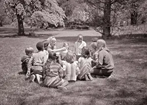 Camp Prints: 1960s ADULT WOMAN TEACHER HANDING A BALL TO ETHNICALLY MIXED GROUP OF CHILDREN VARIOUS AGES SITTING IN CIRCLE ON THE