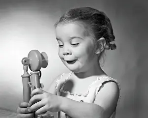 Communications Prints: 1960s LITTLE GIRL WITH FUNNY FACIAL EXPRESSION HOLDING A TOY CANDLE STICK TELEPHONE IN BOTH HANDS