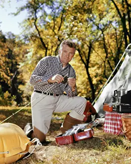 Oldtime Prints: 1960s MAN LOOKING AT CAMERA AT CAMPSITE ATTACHING LURE TO FISHING ROD