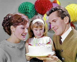 Mid Adultman Prints: 1960s SMILING MOTHER AND FATHER WITH DAUGHTER READY TO BLOW OUT CANDLES ON BIRTHDAY CAKE PARTY DECORATIONS