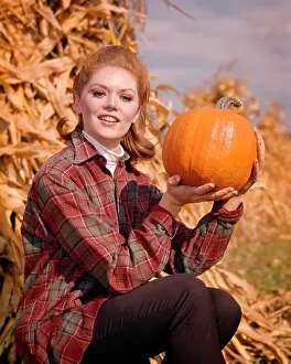 Decision Prints: 1960s SMILING RED HAIR WOMAN WEARING A PLAID SHIRT SITTING BY CORN SHOCK LOOKING AT CAMERA HOLDING UP PUMPKIN