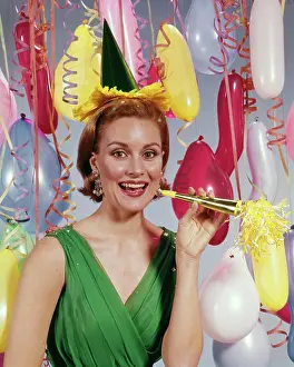 Oldtime Prints: 1960s SMILING WOMAN LOOKING AT CAMERA AMID STREAMERS AND BALLOONS HOLDING NEW YEARS NOISE MAKER