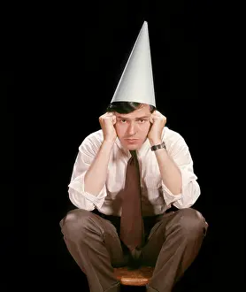 Oldtime Prints: 1960s UNHAPPY YOUNG MAN SITTING ON STOOL WEARING DUNCE CAP LOOKING AT CAMERA