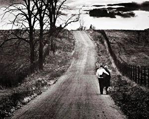 Dreams Prints: 1960s BACK VIEW MATURE COUPLE ARM IN ARM WALKING DOWN LONELY ROAD BARREN AUTUMN LANDSCAPE WITH OMINOUS CLOUDS ON
