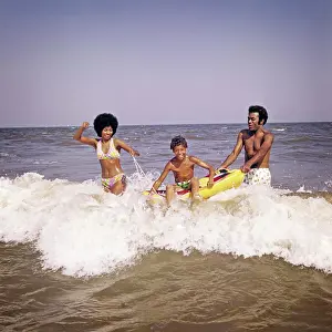 Dads Prints: 1970s AFRICAN-AMERICAN FAMILY JUMPING IN OCEAN SEASHORE SURF MOTHER AND FATHER WITH SON ON INFLATABLE RAFT