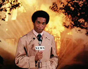 Grownup Prints: 1970s AFRICAN AMERICAN MAN TELEVISION NEWS REPORTER WEARING RAINCOAT SPEAKING INTO MICROPHONE AT FIRE SCENE LOOKING AT