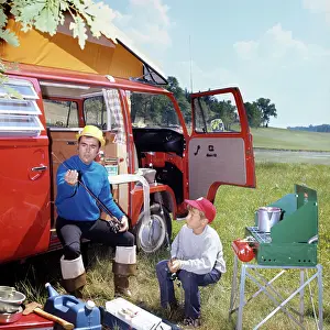 Mid Adultman Prints: 1970s MAN AND BOY CAMPING SON SITTING BY PORTABLE STOVE FATHER SITTING BY CAMPER WITH FISHING RODS AND GEAR