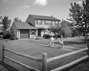 Best Prints: 1970s TWO YOUNG GIRLS SISTERS RIDING BICYCLES IN YARD OF SUBURBAN HOUSE