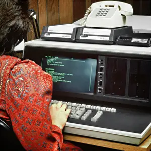 Anonymous Prints: 1980s BACK OF ANONYMOUS WOMAN WEARING RED PAISLEY DRESS WORKING ON EARLY COMPUTER TERMINAL KEYBOARD WITH DIAL-UP