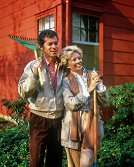 Expressions Prints: 1980s MATURE COUPLE STANDING BY HOUSE MAN HOLDING RAKE ON SHOULDER WOMAN LEANING ON GARDEN TOOL WEARING AUTUMN CLOTHES