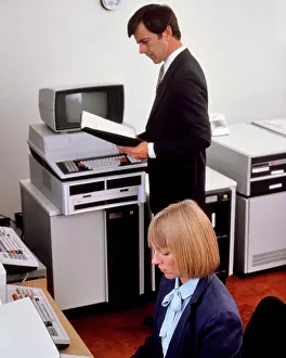Innovation Prints: 1980s OFFICE WITH EARLY COMPUTER HARDWARE WOMAN SEATED AT TERMINAL KEYBOARD MAN STANDING READING MANUAL