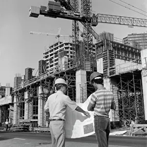 Manly Prints: 1980S Rear View Of Two Hard Hat Workers Standing In Front Of Major Construction Site Looking Over Building Plans Men