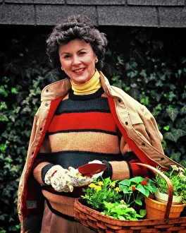 Sweater Prints: 1980s SMILING WOMAN WEARING SWEATER JACKET YELLOW TURTLENECK GARDENING GLOVES HOLDING BASKET OF PLANTINGS LOOKING AT