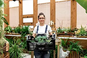 Carry Prints: Adult happy woman smiling looking at camera and carrying plastic box with succulents while working in wooden greenhouse