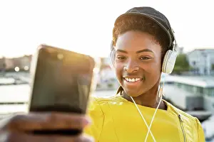 Balcony Prints: African American woman in stylish bright jacket using mobile phone and listen to music on headphones