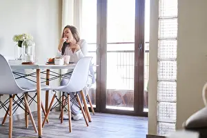 Balcony Prints: Attractive young happy woman having breakfast at table near window at home