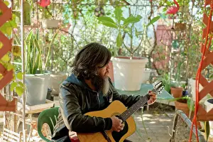 Greenery Prints: A bearded man with long hair plays an acoustic guitar in a cozy garden surrounded by potted plants and decorative items