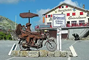 Mountain Chain Prints: Biker couple, metal sculpture on the Grimsel Pass in front of the Berghotel Alpenlodge Grimselpass
