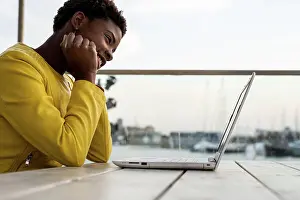 Attractive Prints: Black African American woman in yellow jacket using laptop at wooden desk in city on blurred background