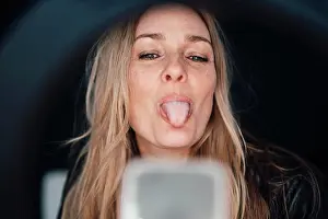 45 To 50 Years Prints: Blond mature influencer sticking tongue out vlogging through smart phone