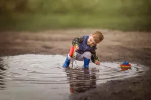 1 2 Years Old Prints: Boy in rain boots crouching in puddle and playing with ships