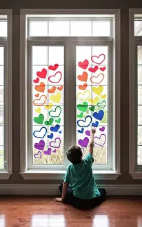 Gradeschooler Prints: Boy sitting on floor by a window covered in a rainbow of paper hearts
