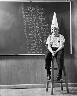 One Child Only Prints: Boy wearing a dunce cap sitting in front of a blackboard