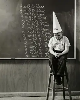 Blackboard Prints: Boy wearing a dunce cap sitting in front of a blackboard