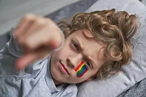 Cheerful Prints: Cheerful boy with rainbow under eye smiling and pointing at camera while lying on blanket