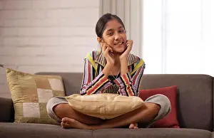 Preteens Prints: Cheerful girl sitting on sofa and looking at camera with hands on cheek