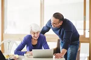 Computers Prints: Cheerful mature man standing by mother looking at laptop on table in nursing home