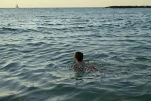 Sailboat Prints: Child swimming in the sea at sunset with a distant sailboat on the horizon. Israel