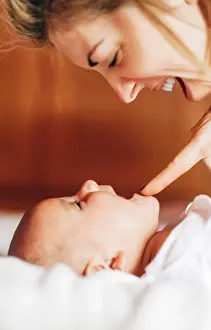 Closeup of glad mom touching nose by nose of baby playing with newborn infant with open mouth having fun lying on bed