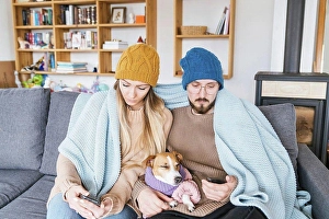 Austerity Prints: Couple with dog sitting on couch wrapped in blanket looking at smartphone