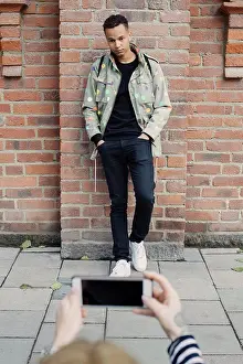 Brickwall Prints: Cropped hands of young woman photographing fashionable man standing with hands in pockets against brick wall
