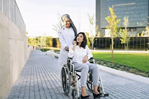 Disability Prints: Disabled woman talking with female doctor pushing wheelchair on footpath