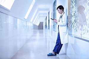 Consumer Electronics Prints: Doctor in hospital corridor leaning against wall using smartphone