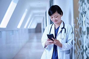 Consumer Electronics Prints: Doctor in hospital corridor leaning against wall using smartphone